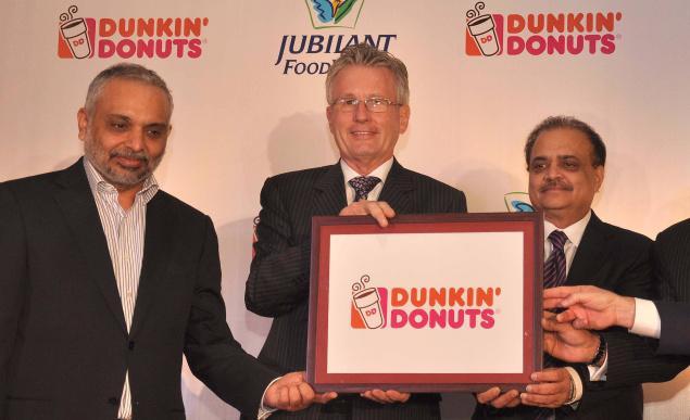 The image shows the official launch event of Dunkin' Donuts in India, following a master franchise agreement signed with Jubilant FoodWorks Limited. Three key executives are pictured holding a framed Dunkin' Donuts logo: • Left: Dr. Amit Varma, who served as a Non-Executive Director of Jubilant FoodWorks. • Center: Nigel Travis, who was the Chief Executive Officer and President of Dunkin' Brands. • Right: Shyam S. Bhartia, the Chairman and Director of Jubilant Bhartia Group